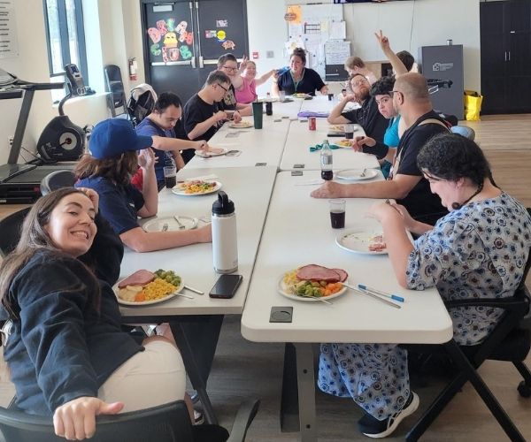 A group of people sit around a long table, sharing a meal and smiling. Plates of food and drinks are in front of them. Some are engaging with each other while one person in front smiles at the camera.