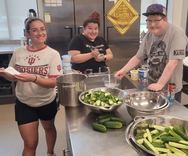 Three people are smiling and preparing food in a kitchen. They are slicing cucumbers and have bowls, utensils, and ingredients on a metal table. 