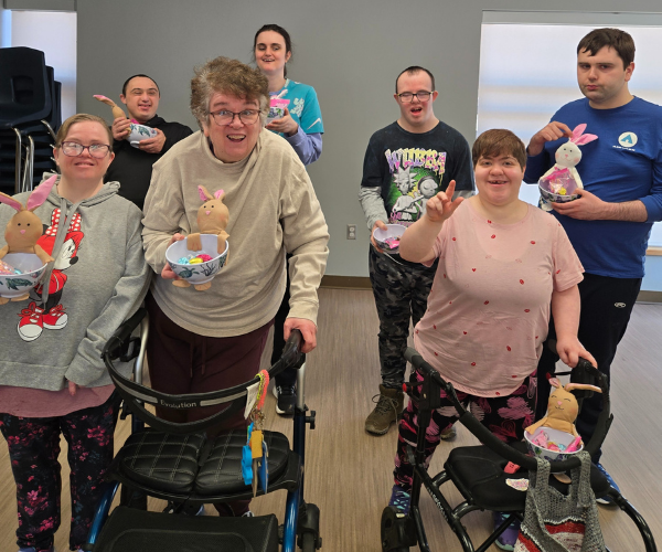 A group of seven smiling adults, some using walkers, hold Easter baskets they made in craft club with stuffed bunnies and eggs, posing together indoors in a bright room.