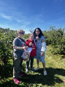 Three women stand together in an apple orchard on a sunny day. One holds a bag of apples. They are smiling and surrounded by green trees under a bright blue sky.