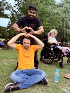 Two men are making a heart shape with their arms; one stands behind the other, who is sitting cross-legged on grass. A woman in a wheelchair is in the background, and there’s a blue water bottle in the foreground.